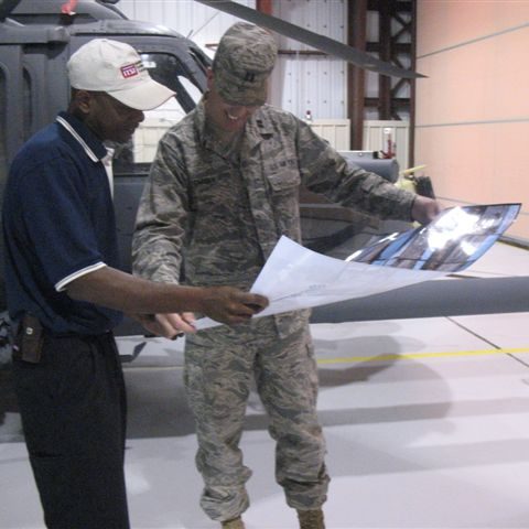 Two men, one in military uniform, examine a document near a helicopter.