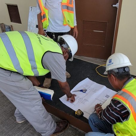 Two workers in safety vests consult blueprints on the ground while a third person observes.